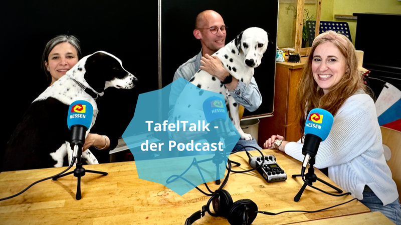 Zwei Lehrkräfte sitzen mit ihren Hunden auf dem Schoß an einem Tisch mit Mikrofonen. Die dirtte Person führt das Interview mit ihnen.